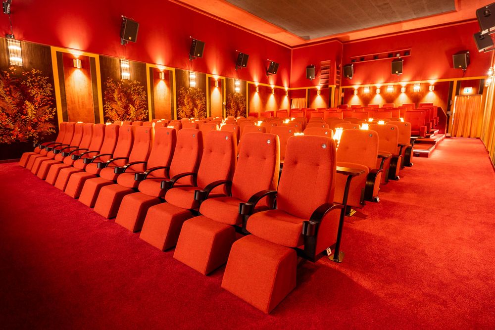 Red-lit interior of a classic movie theater with rows of empty seats and vintage architectural details.