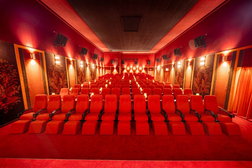 A series of empty red theater seats in a classic movie cinema with dramatic red lighting and ornate wall decor.