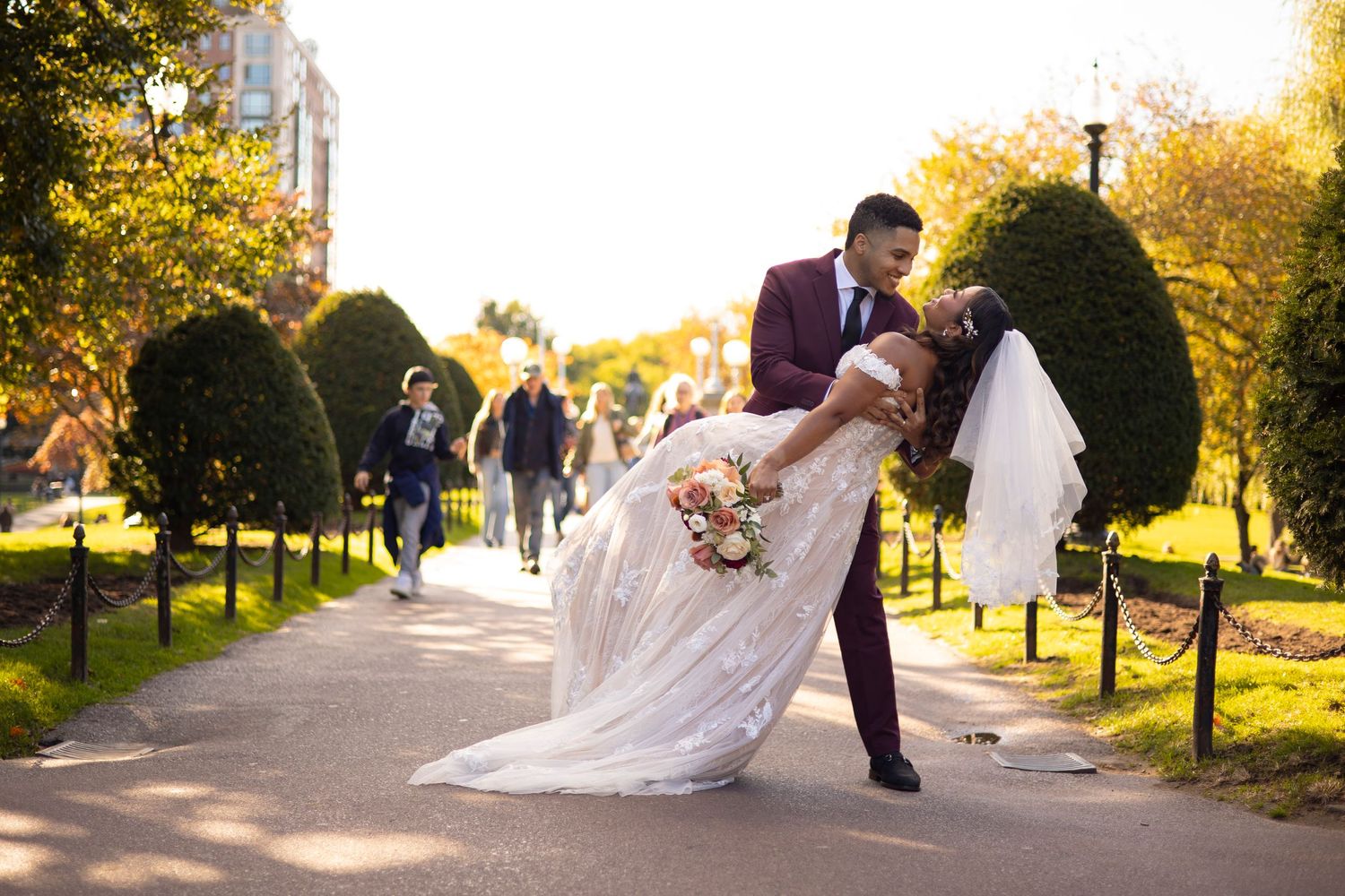 Couple in wedding attire walking along a scenic autumn path with golden sunlight filtering through trees in New England.