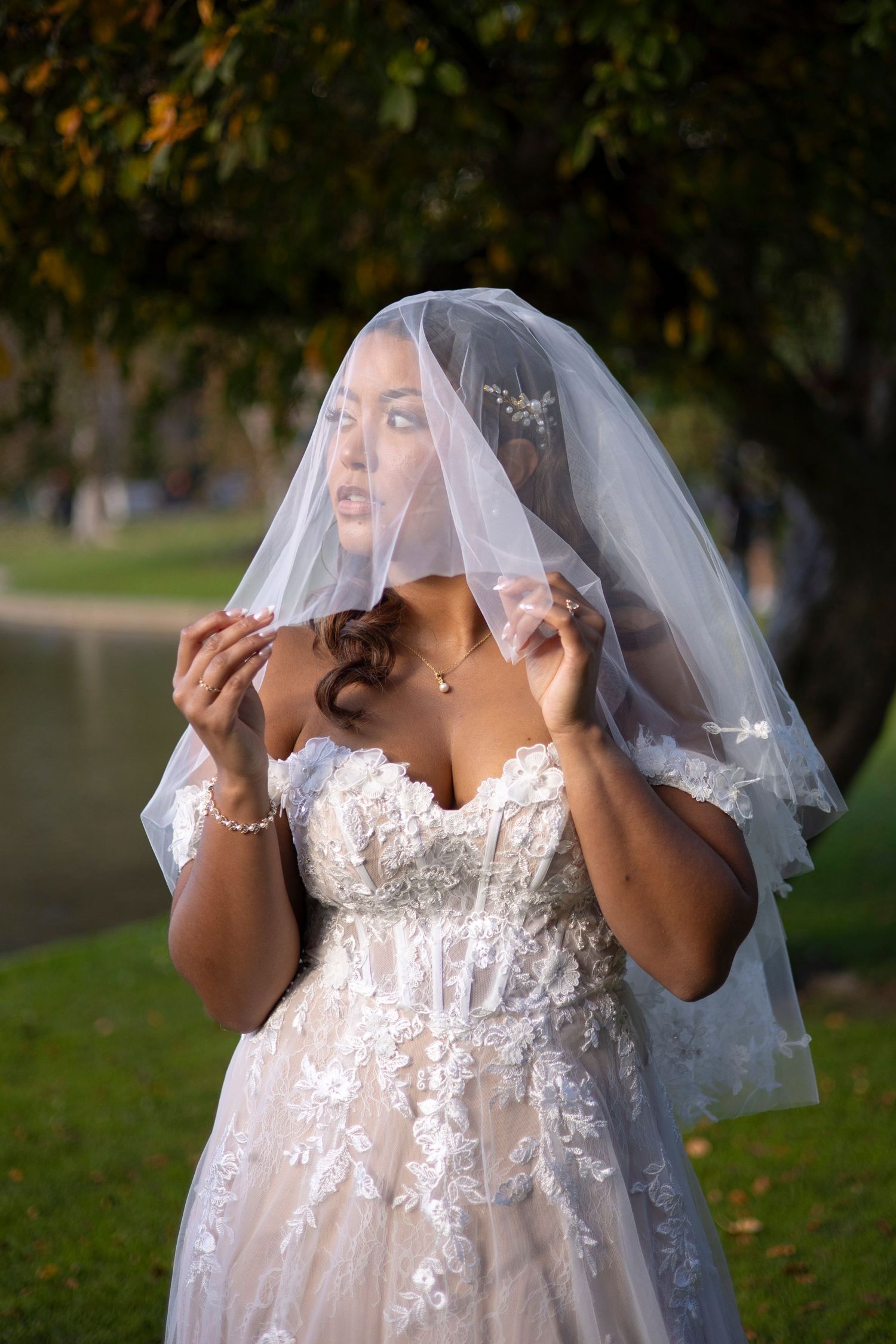 A bride in a white lace dress and veil stands in an outdoor setting with autumn foliage in the background.