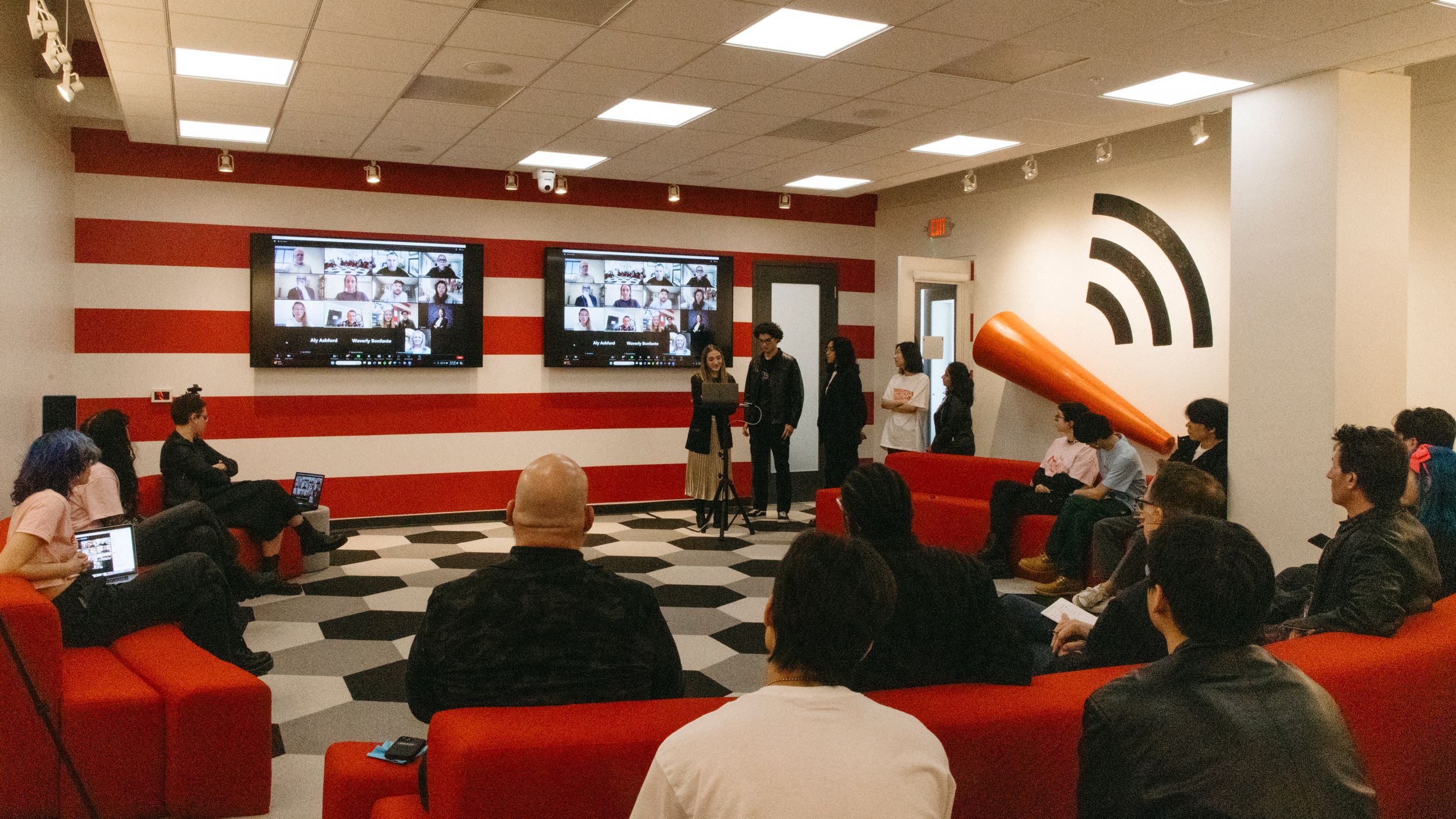 Photo of a presentation room with red stripes on the walls, people presenting, and people watching from couches