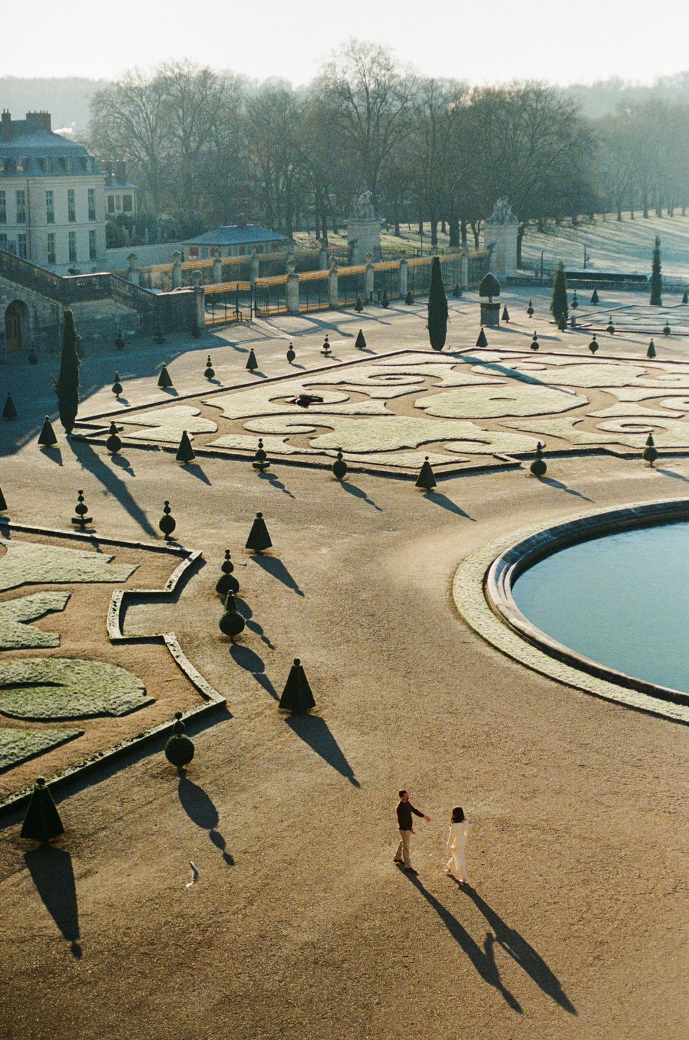 Aerial view of a geometric garden design with long shadows cast by trees and people walking in a park during sunset.