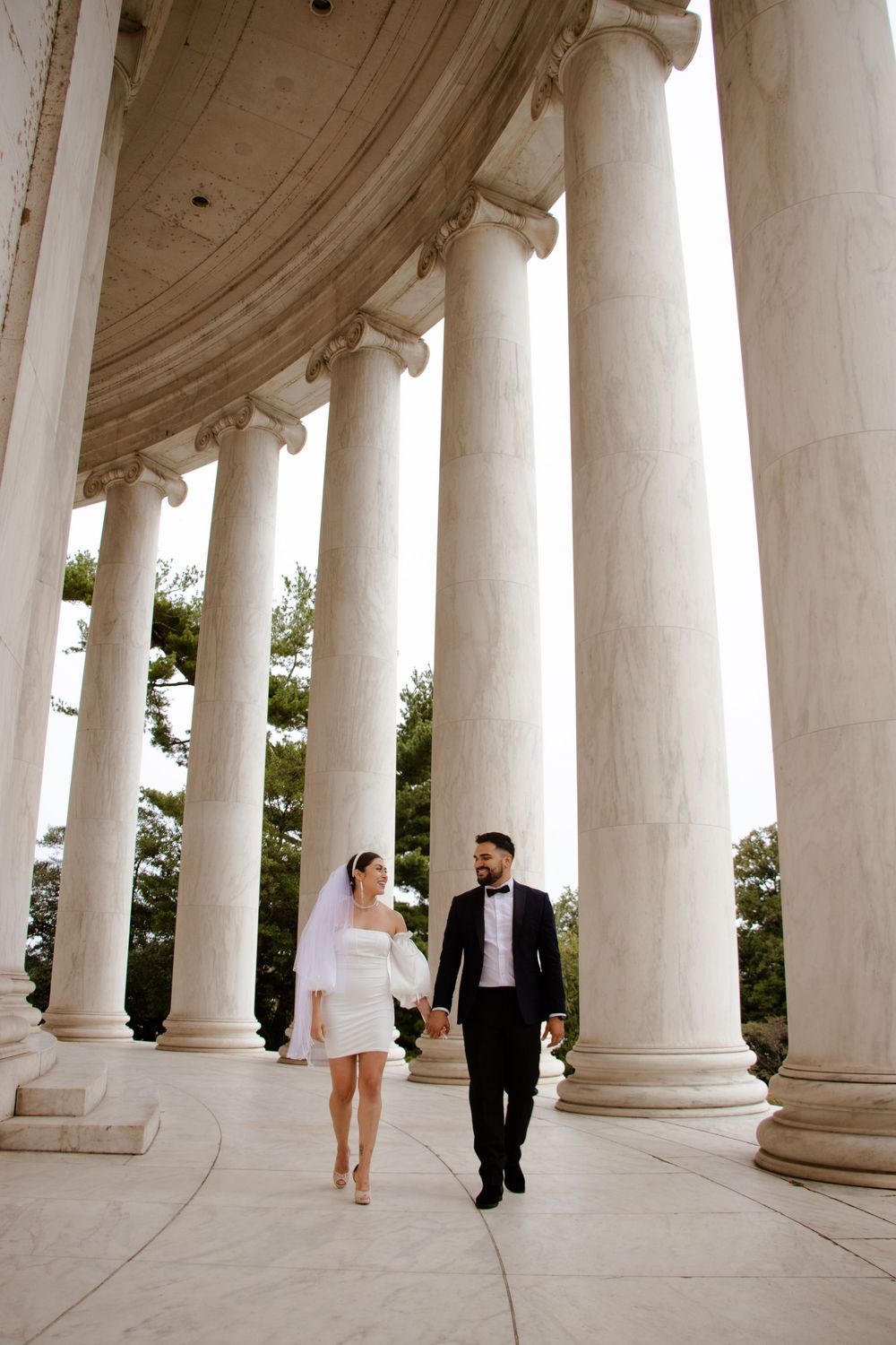 A couple in wedding attire poses between elegant white marble columns at the Jefferson Memorial.