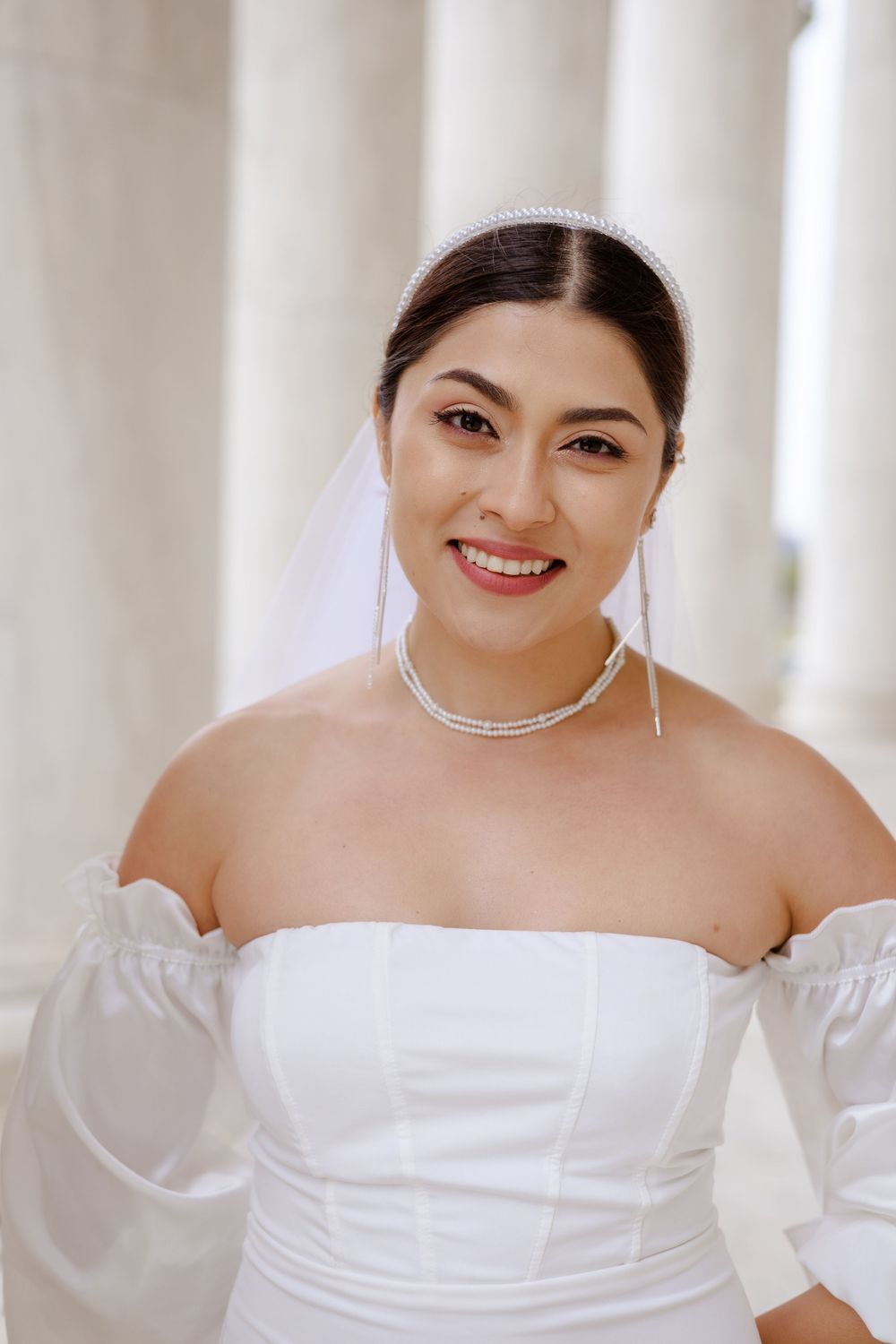 A person in a white off-shoulder dress smiles warmly while wearing a delicate silver necklace.