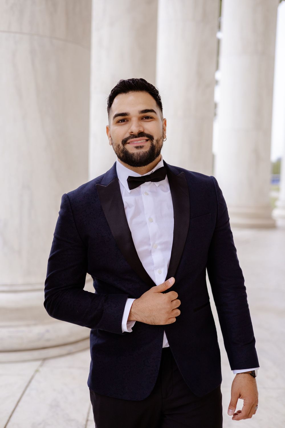 Groom in classic navy tuxedo poses confidently against white pillared backdrop.