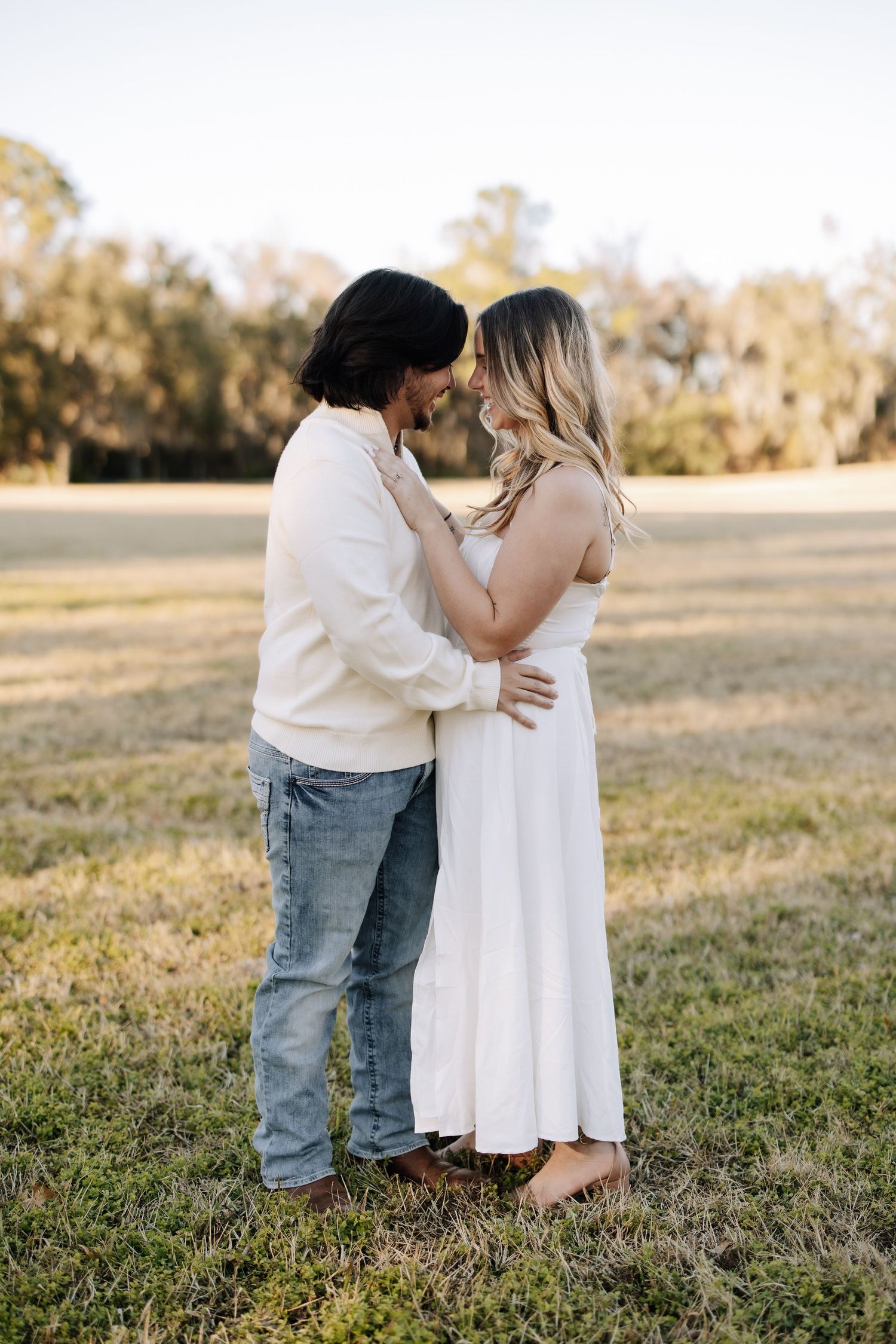 A couple shares a romantic moment sitting in a sun-drenched field at golden hour while dressed in casual summer attire.