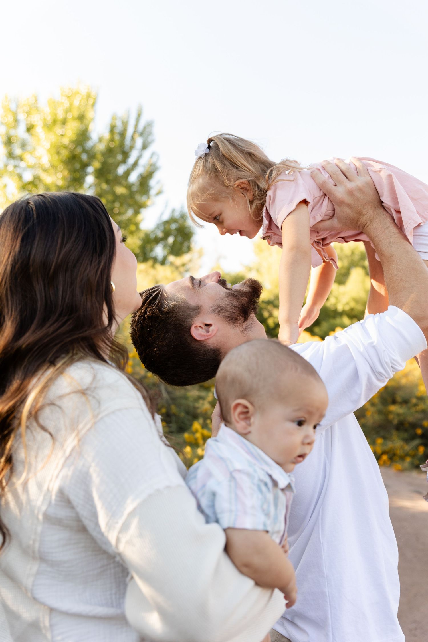 Father lifting laughing toddler girl while mother holds baby boy outdoors in sunny natural setting.