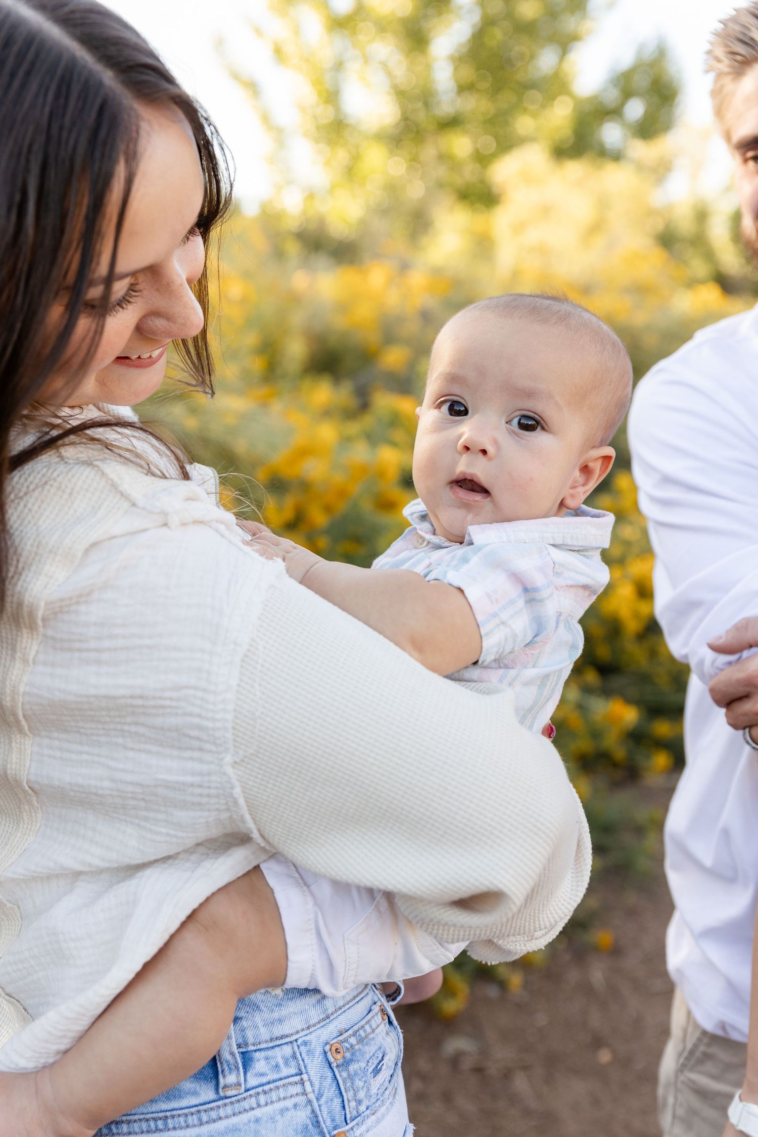 A mother holds her baby outdoors near yellow wildflowers while a man stands beside them on a sunny day.