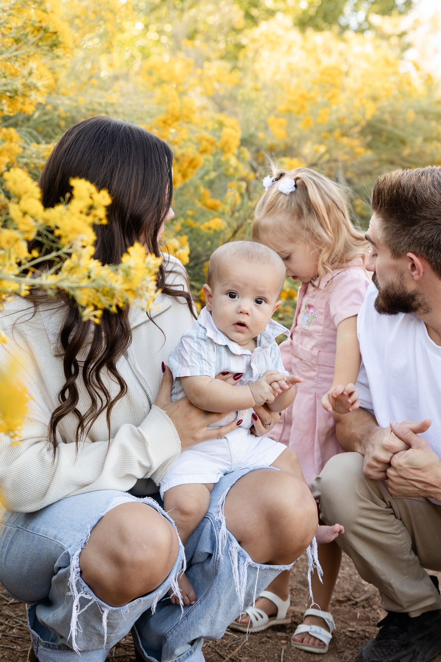 Family with baby and toddler girl crouching outdoors among yellow wildflowers during a warm autumn day.