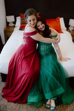 Two people in burgundy and emerald formal dresses sit together on a bed showing off their elegant gowns.