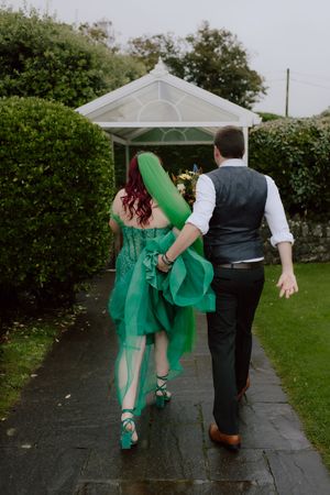 A couple in formal attire playfully dances on a wet garden path, with the woman wearing an emerald green dress.
