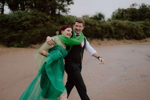 Couple in green dress and black suit share joyful moments during outdoor photo session on sandy path.