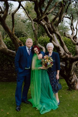 Three people pose together in a garden with twisted trees, with the person in the middle wearing a flowing emerald green dress.