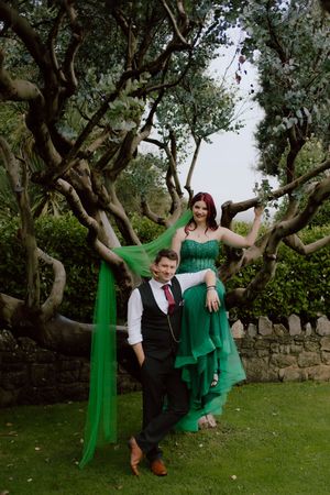 A couple poses together in a garden setting with twisted trees, wearing green and black formal attire.