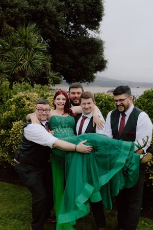 A group in formal attire including a vibrant emerald green dress pose playfully together outdoors against lush greenery.