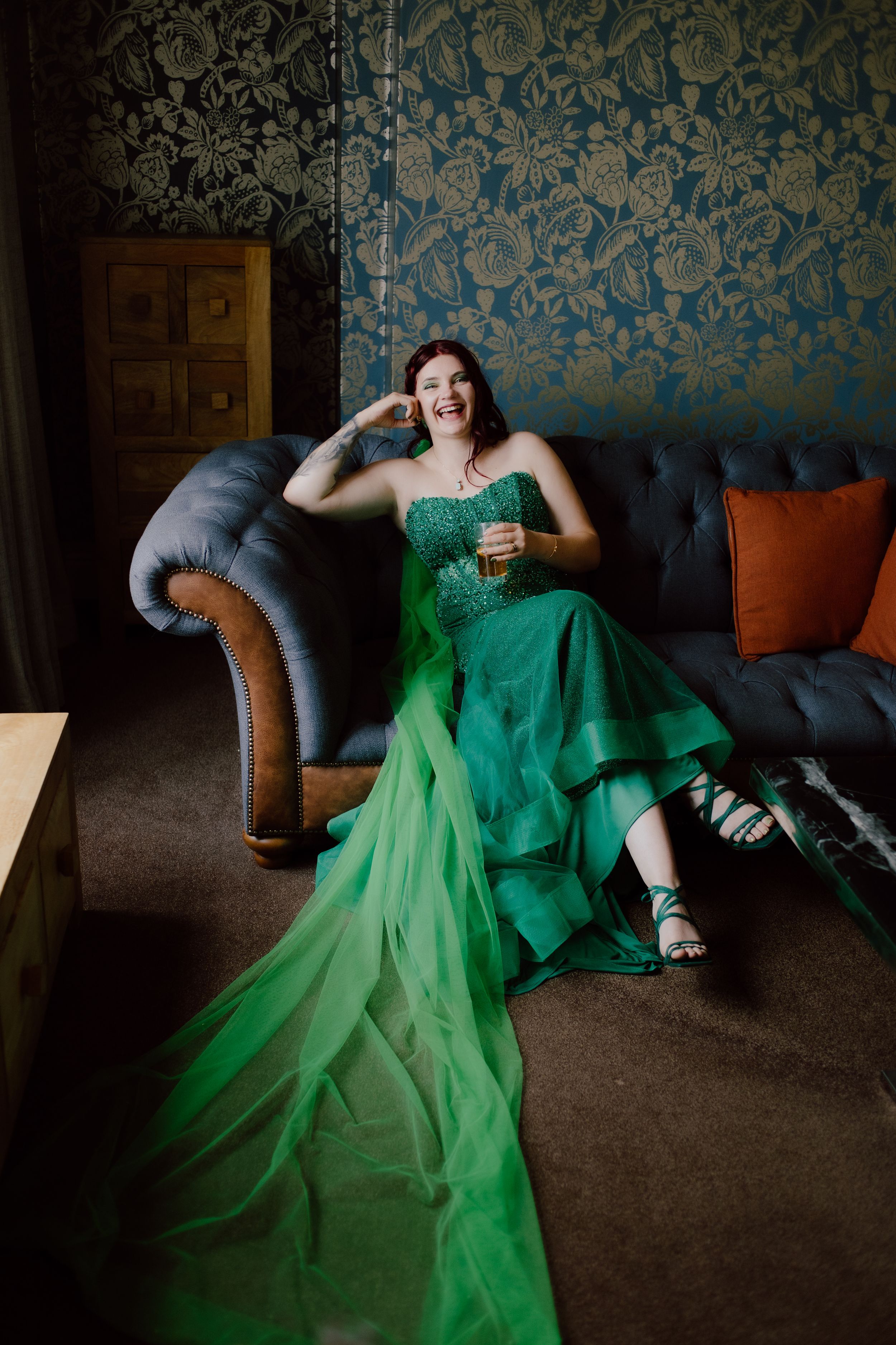A person in a vibrant emerald green evening gown lounges dramatically across a gray couch against blue damask wallpaper.