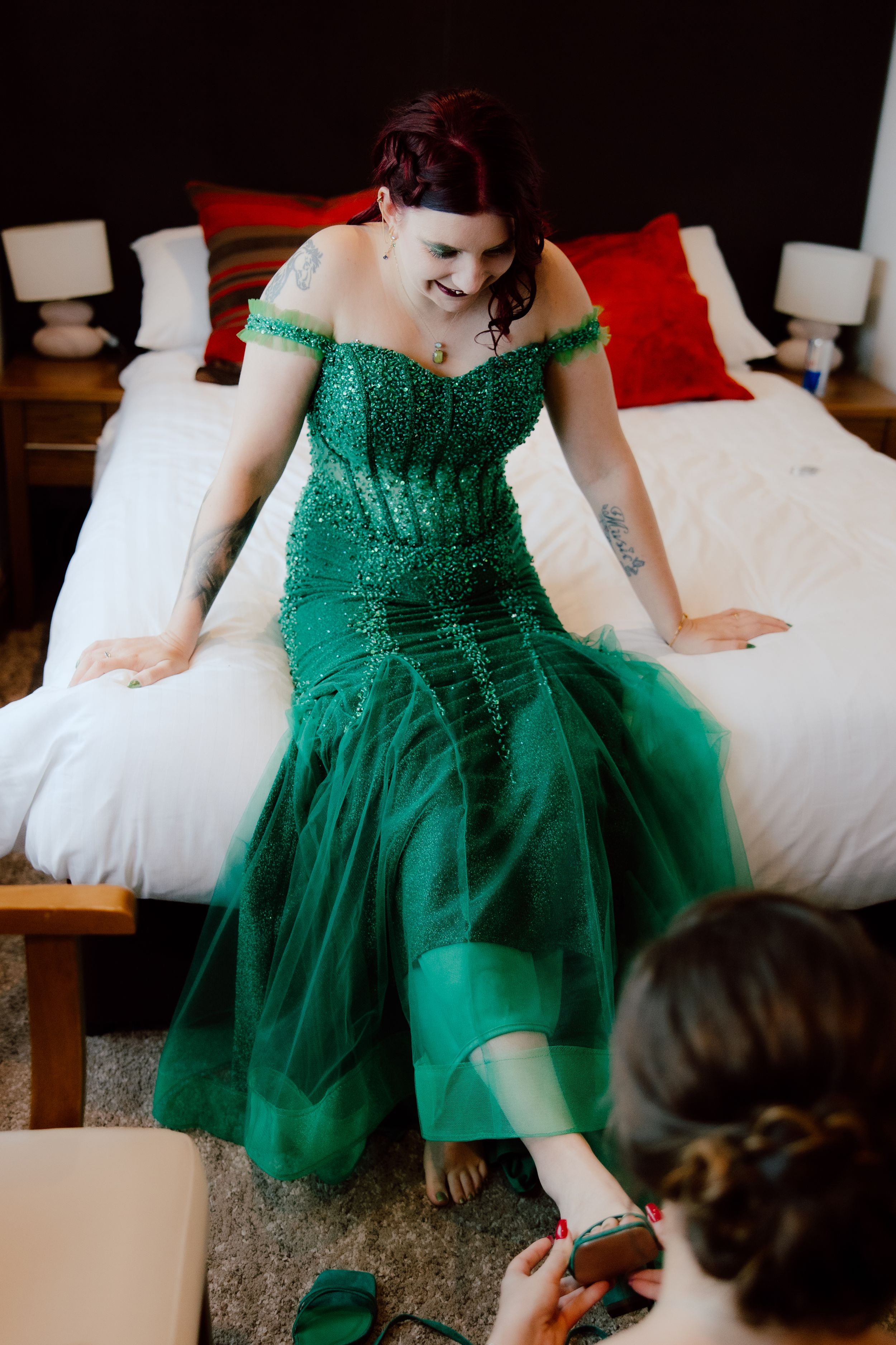 A flowing emerald green tulle gown with off-shoulder detail is displayed on a white bed.
