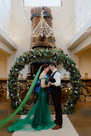 Couple shares romantic moments under a large floral arch with twinkling lights and emerald green wedding attire.