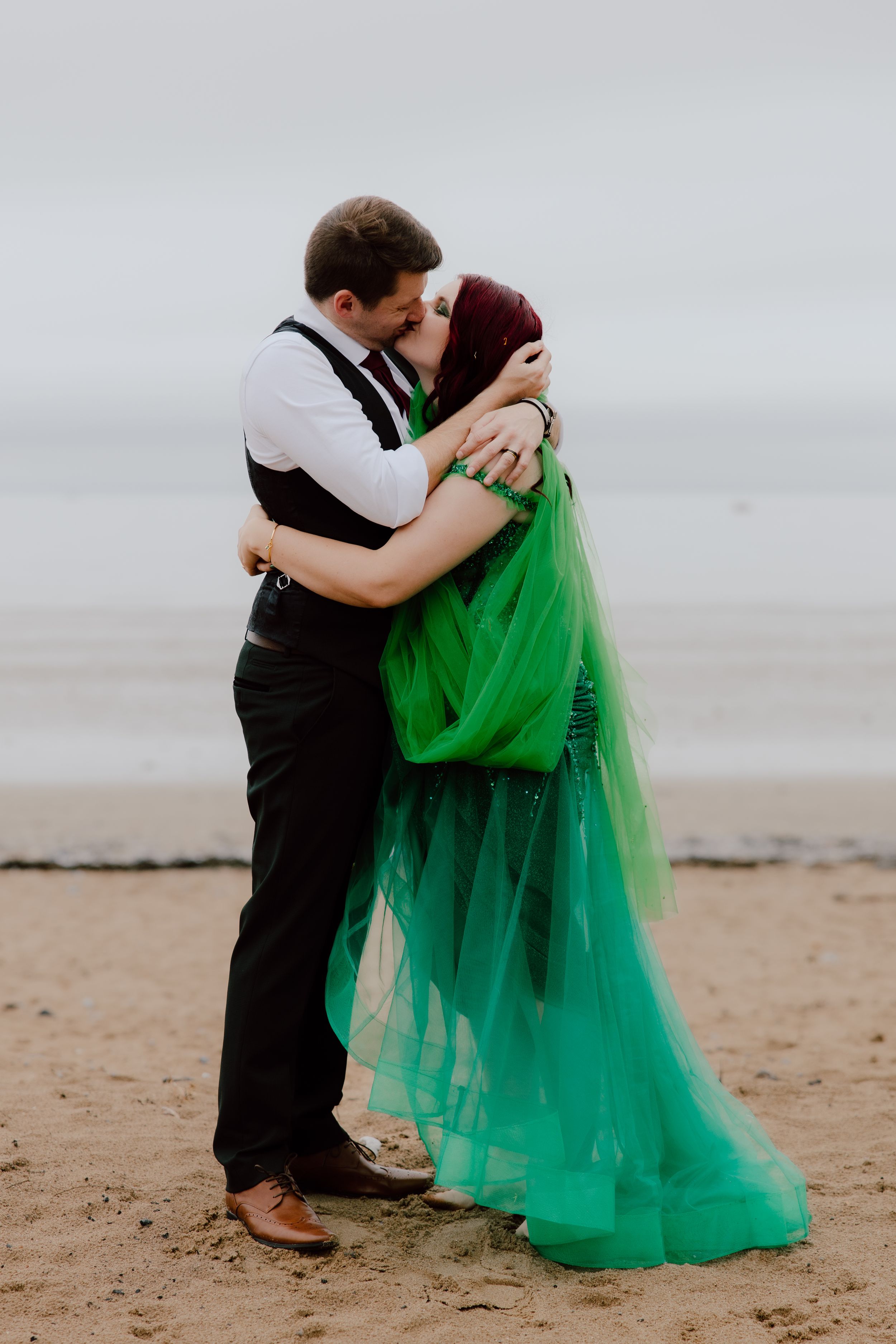A couple embraces on a beach, with one wearing a flowing emerald green tulle dress and the other in black attire.