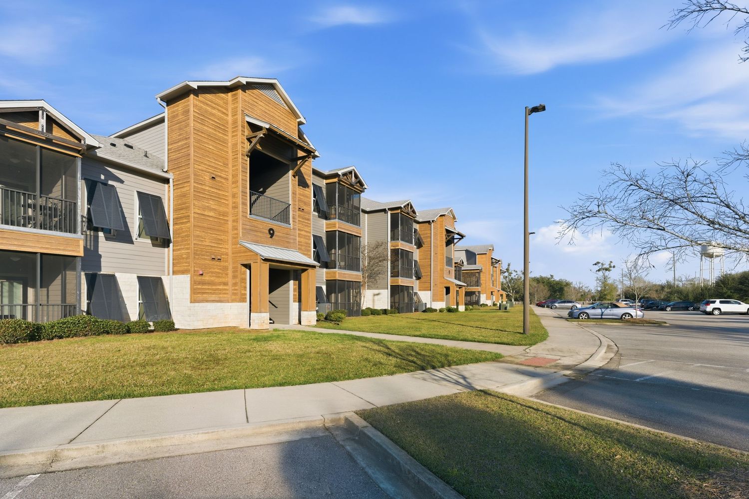 Exterior view of tan brick apartment buildings with parking areas along a suburban street.