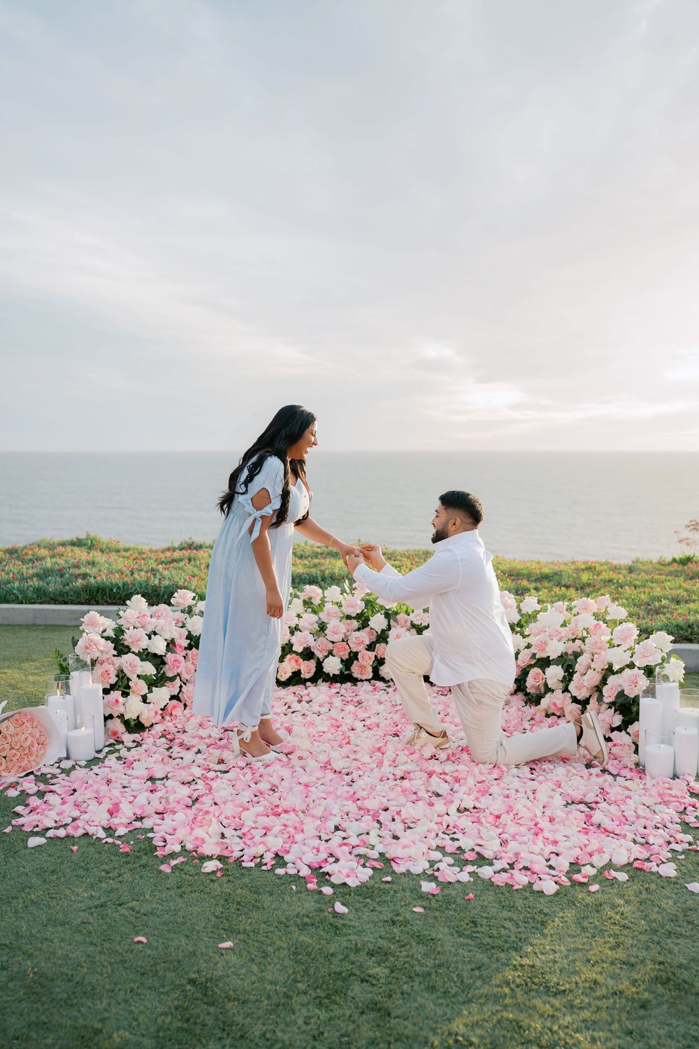 A romantic proposal setup on a hilltop with pink rose petals scattered on grass and white floral arrangements at sunset.