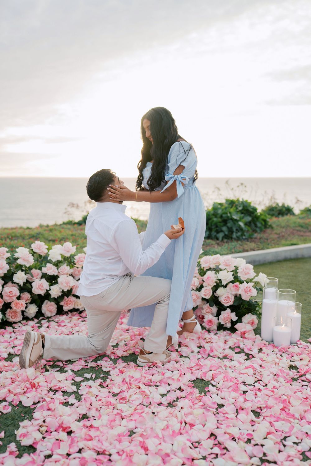 A romantic seaside proposal setup with pink rose petals scattered on the ground and white candles creating an intimate atmosphere.