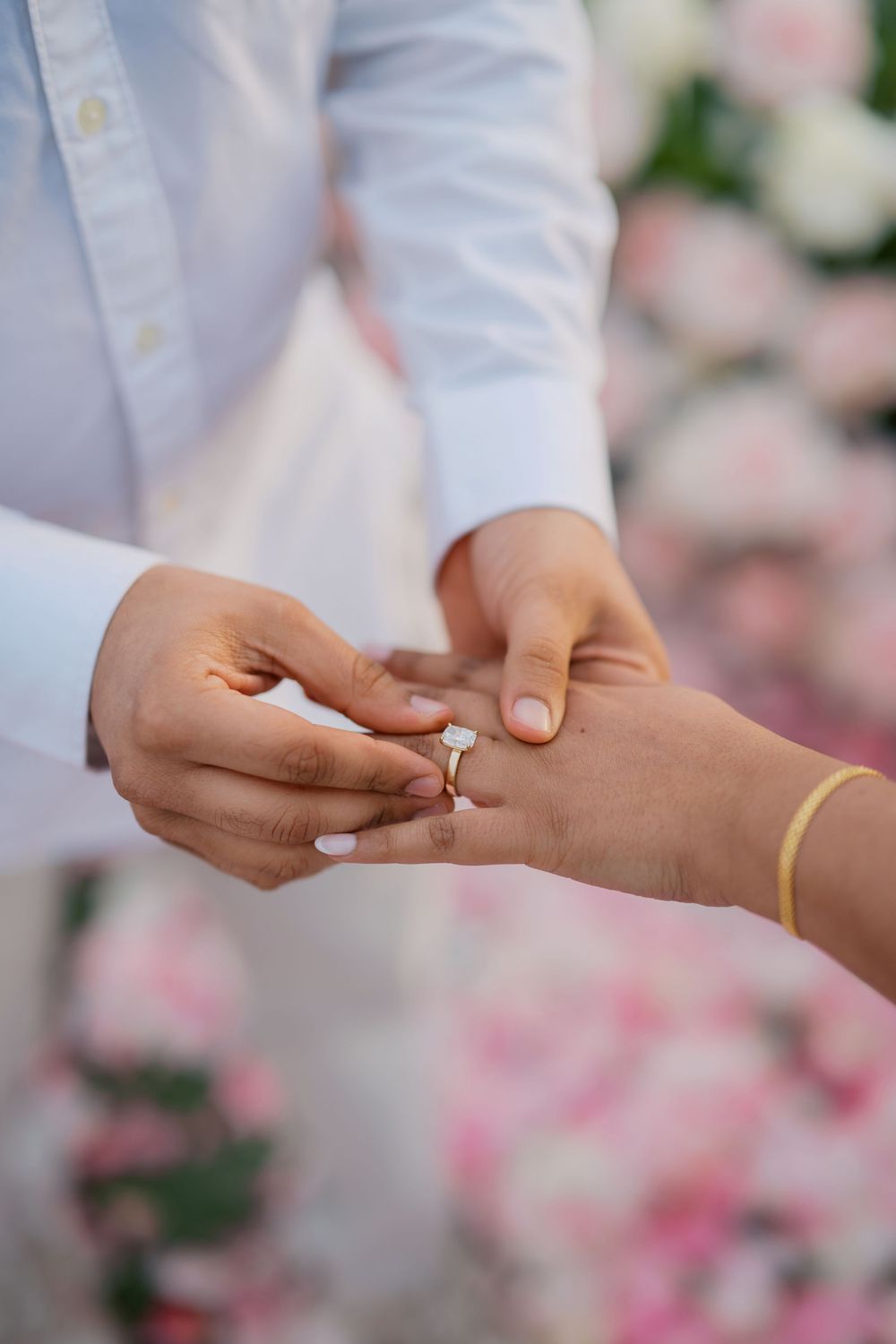 A close-up sequence of hands exchanging and placing a wedding ring during a romantic ceremony with pink floral backdrop.