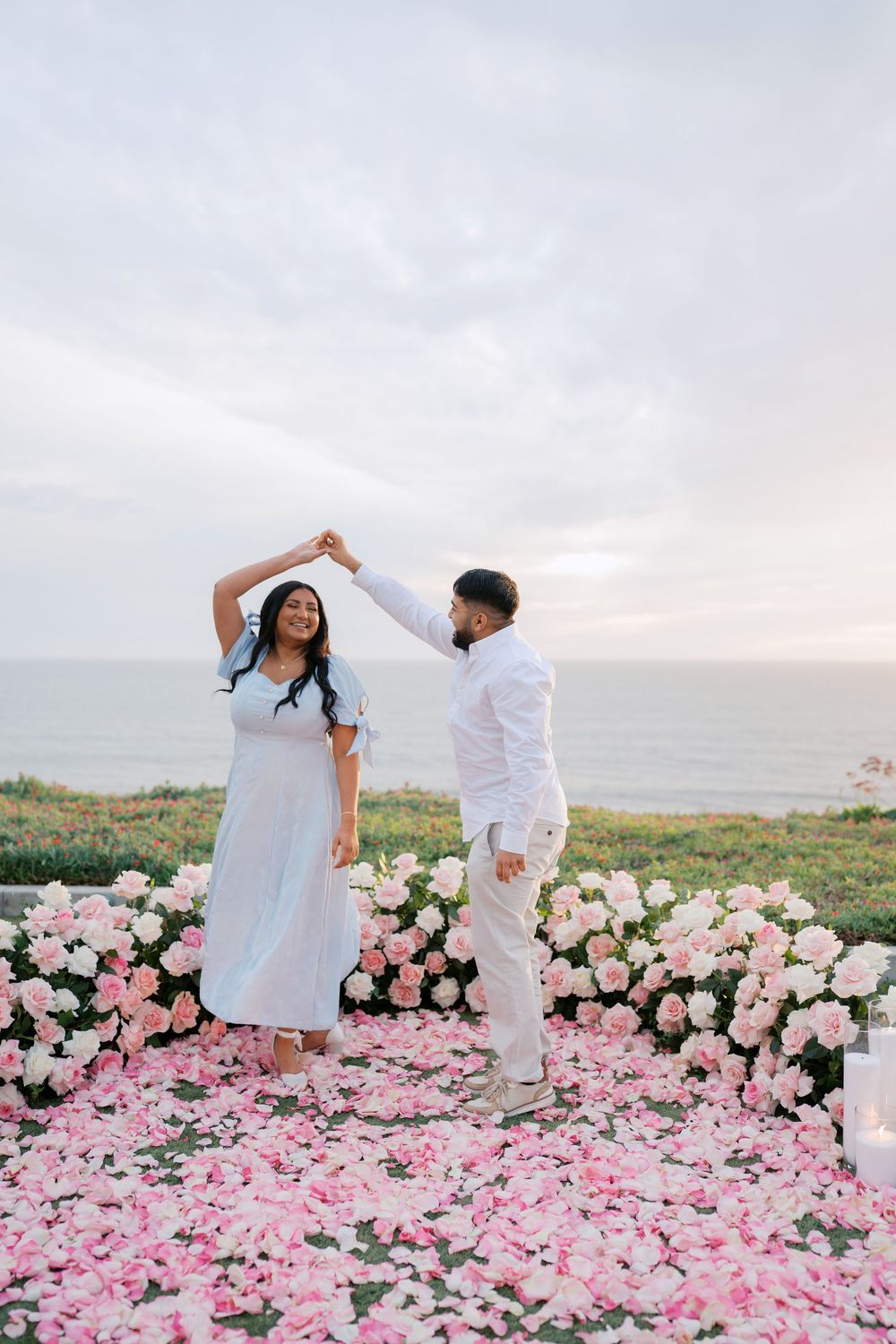 A couple in white outfits dances together in a field of pink and white roses during sunset.
