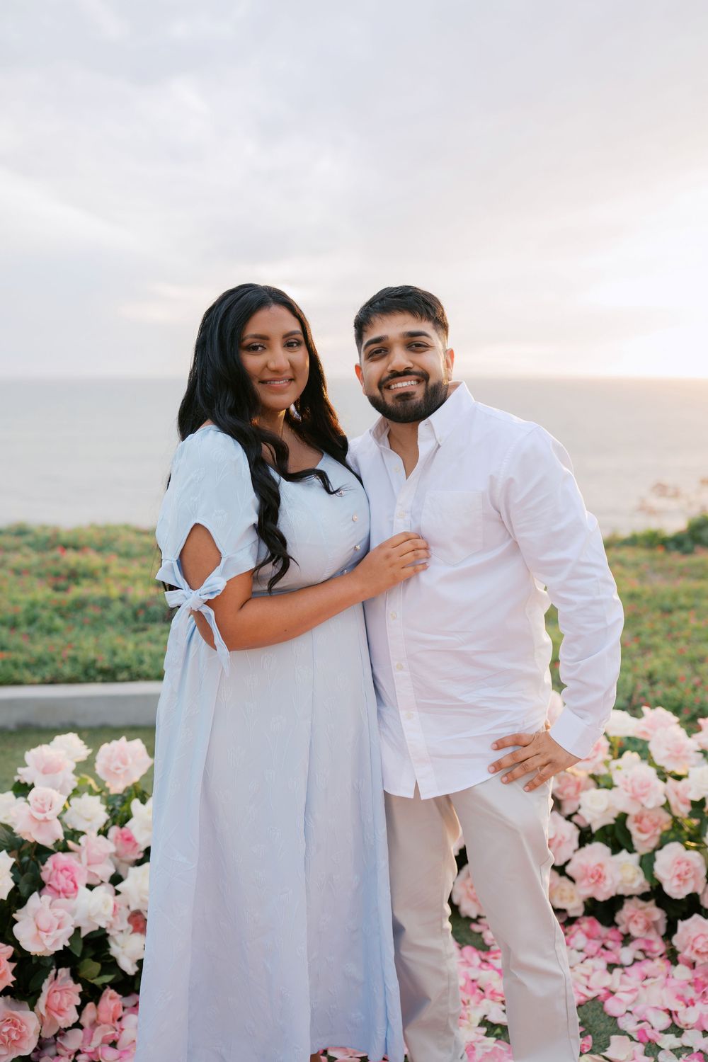 A romantic couple in white outfits poses together near pink roses during a sunset photoshoot.