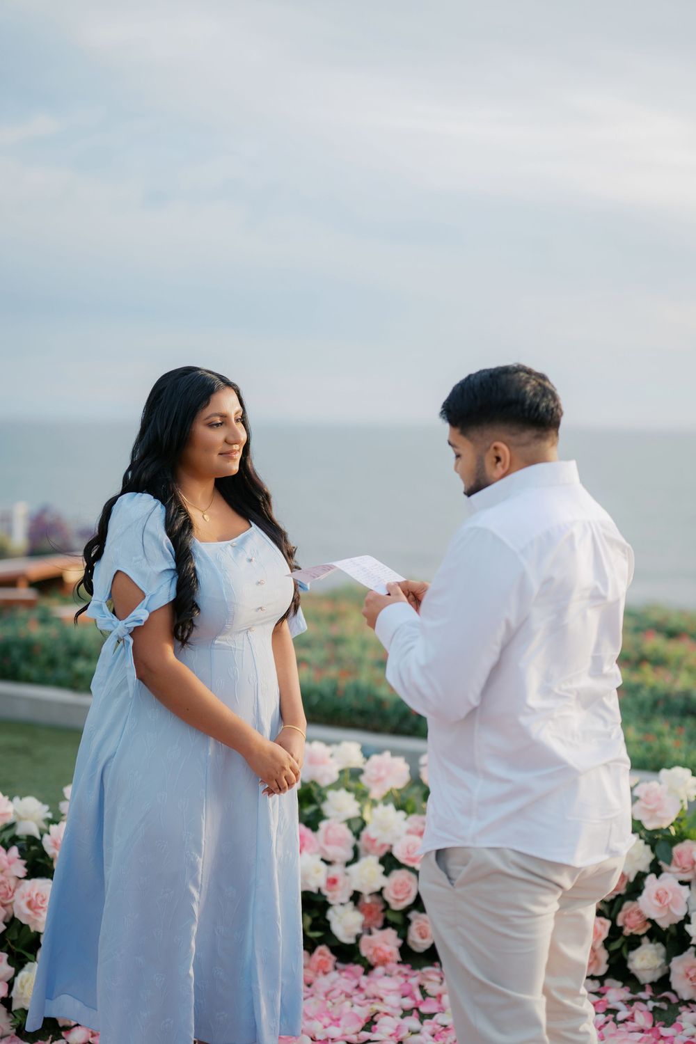 A romantic proposal scene with pink roses and ocean backdrop as a couple shares a special moment in white and blue attire.