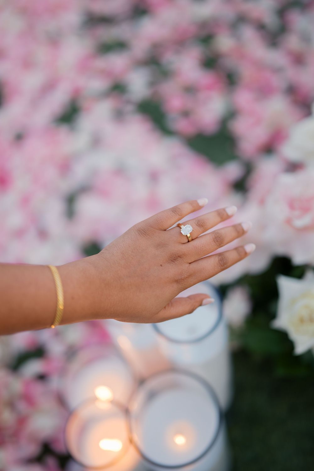 A hand wearing gold rings is shown against a soft pink floral backdrop with glowing candles creating romantic ambiance.