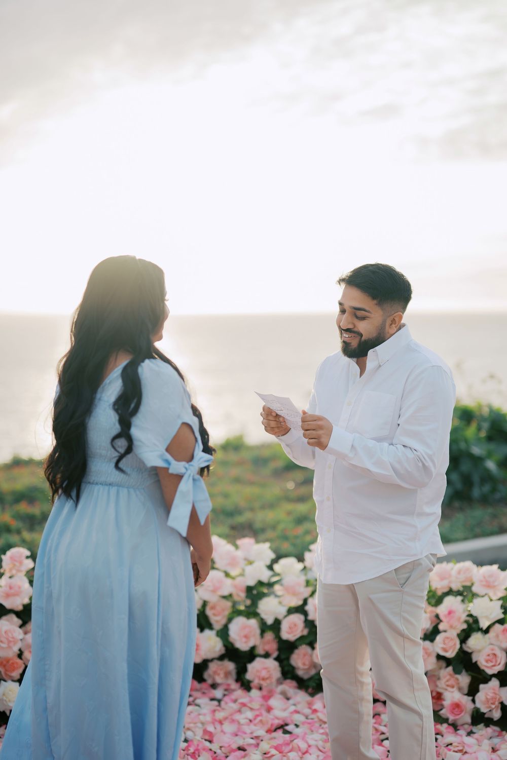 A romantic proposal scene at sunset with pink roses and a couple in white and light blue attire.