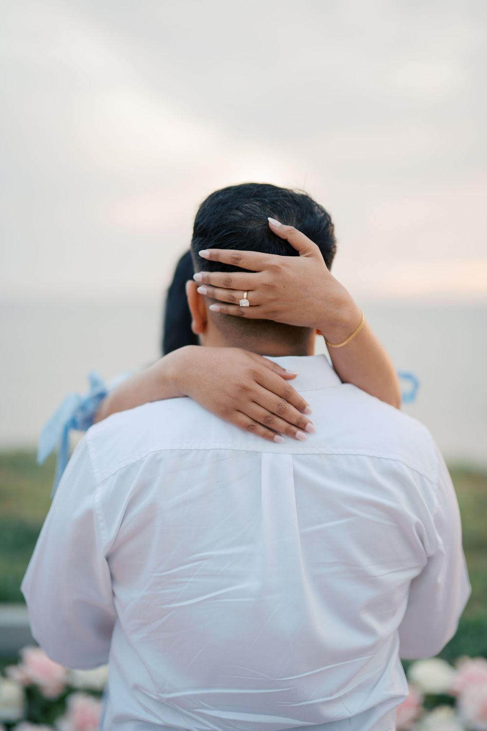 A tender moment captured from behind as hands embrace in white attire against a soft sky.