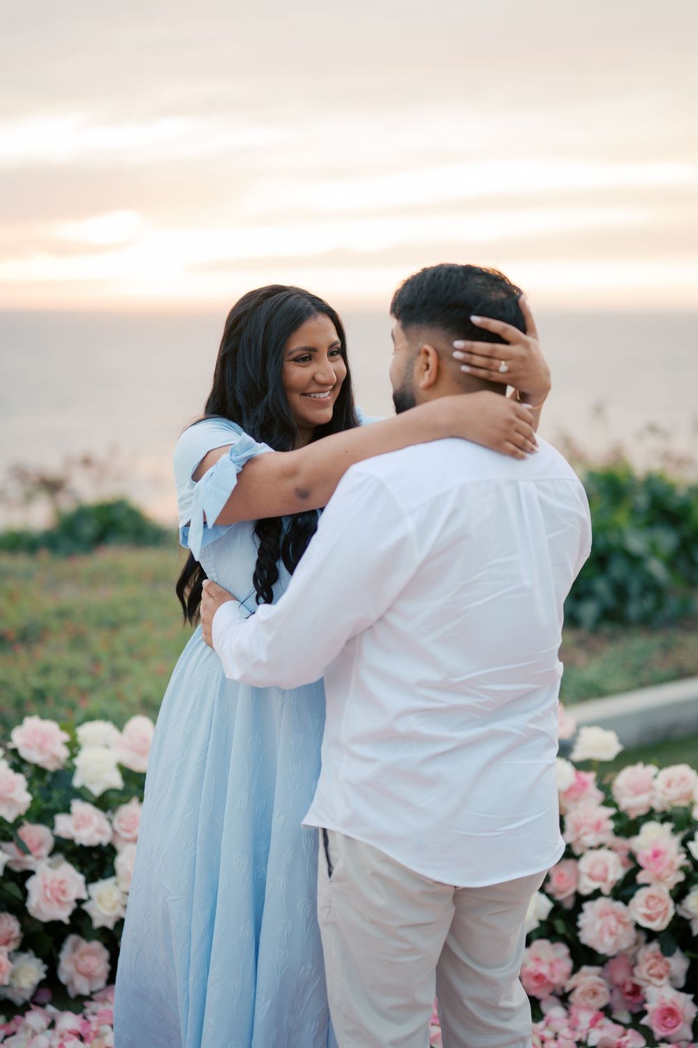 A romantic couple embraces at sunset surrounded by pink roses in a coastal setting.
