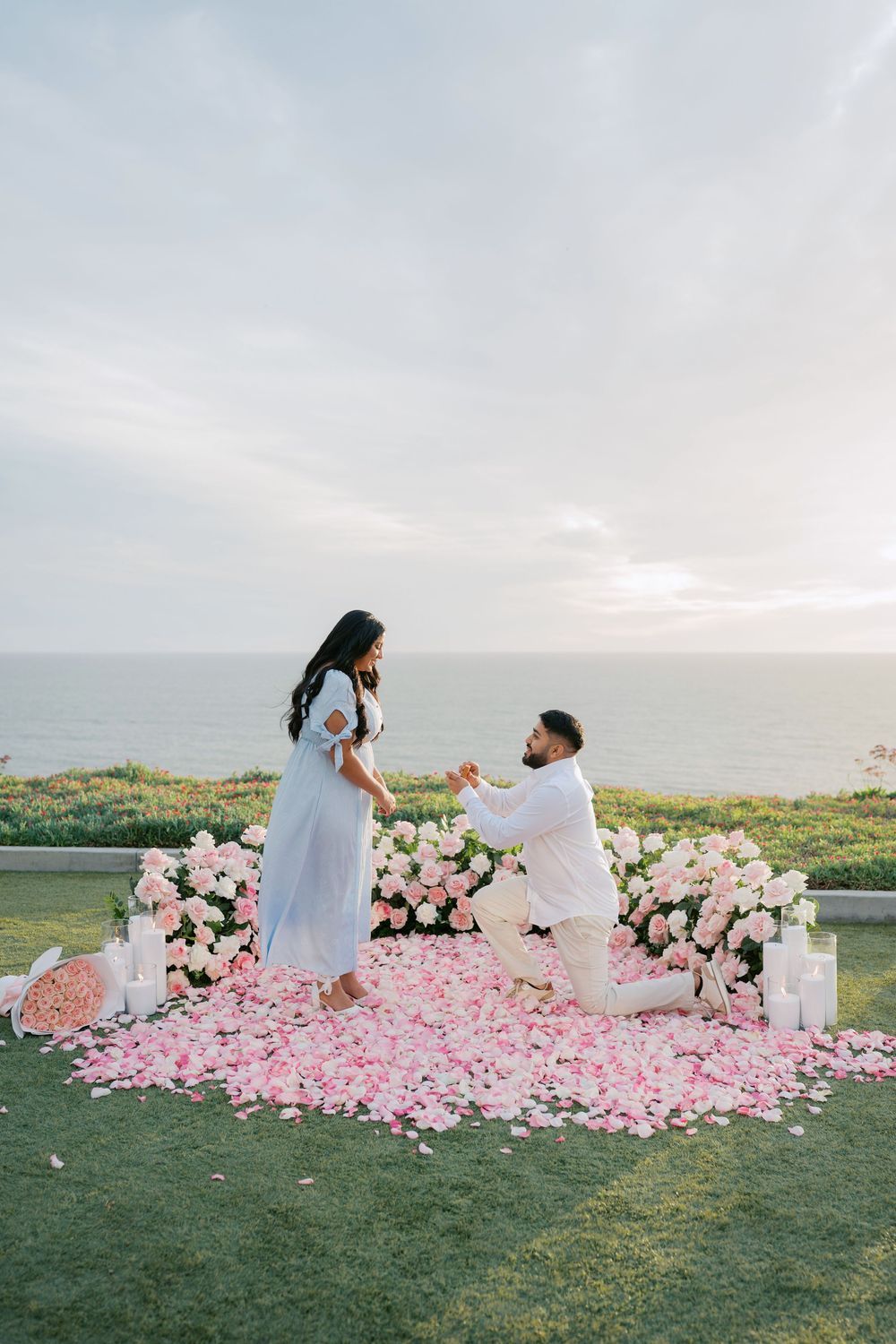 A romantic proposal setup on a grassy hilltop with pink rose petals and white floral arrangements at sunset.