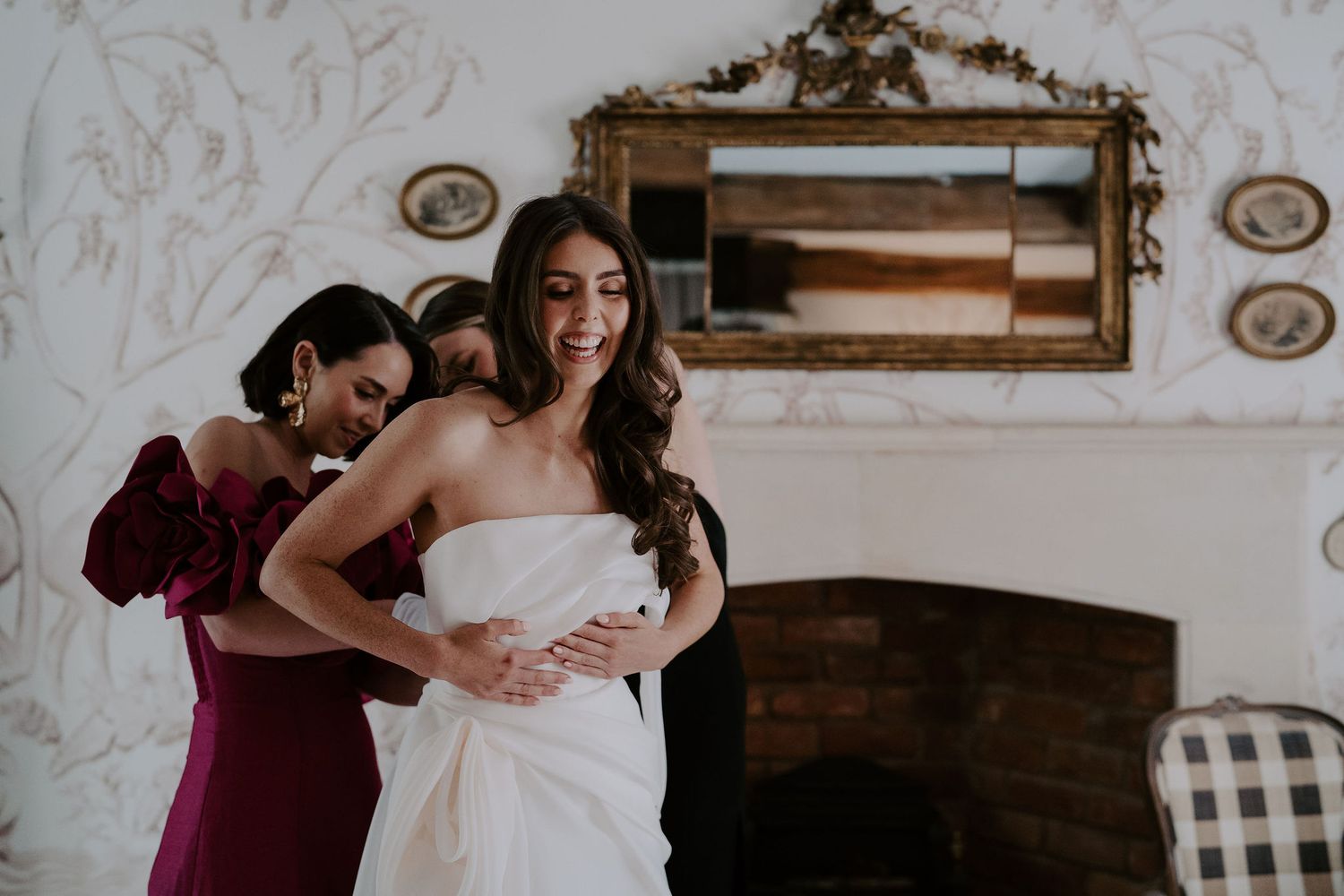 brides sister and friend fastening wedding dress as bride laughs at dewsall court in herefordshire