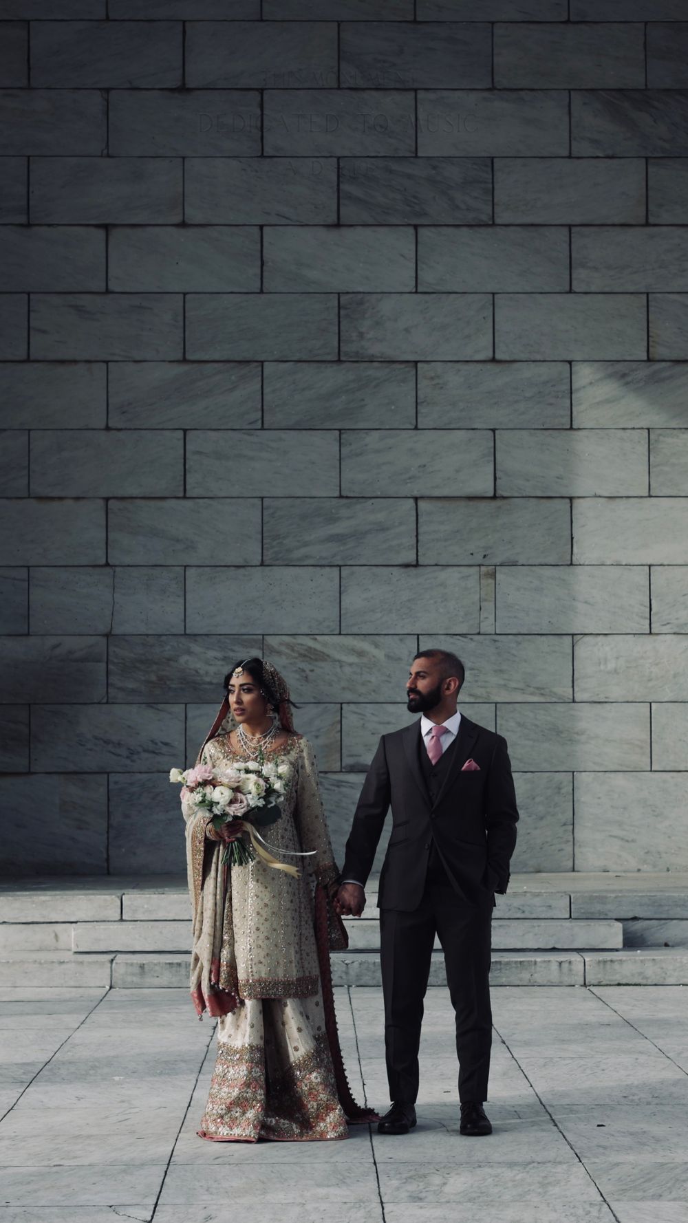South Asian bride in gold lehenga holds bouquet with groom in dark suit, posing before a marble stone wall.