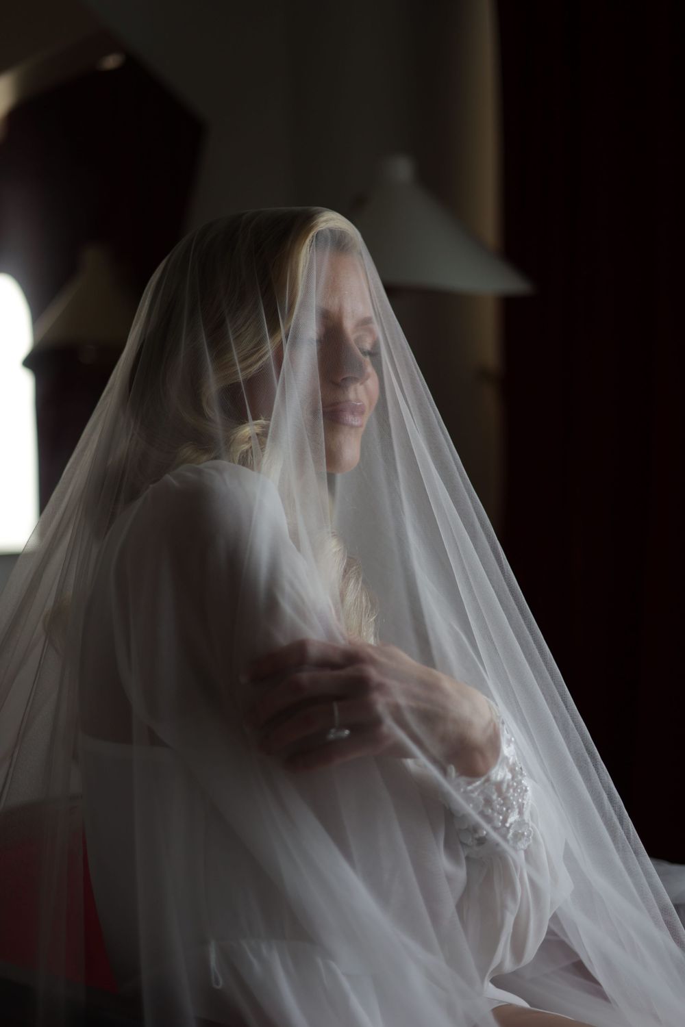 Bride draped in sheer white veil, sitting peacefully in soft light wearing lace wedding gown.
