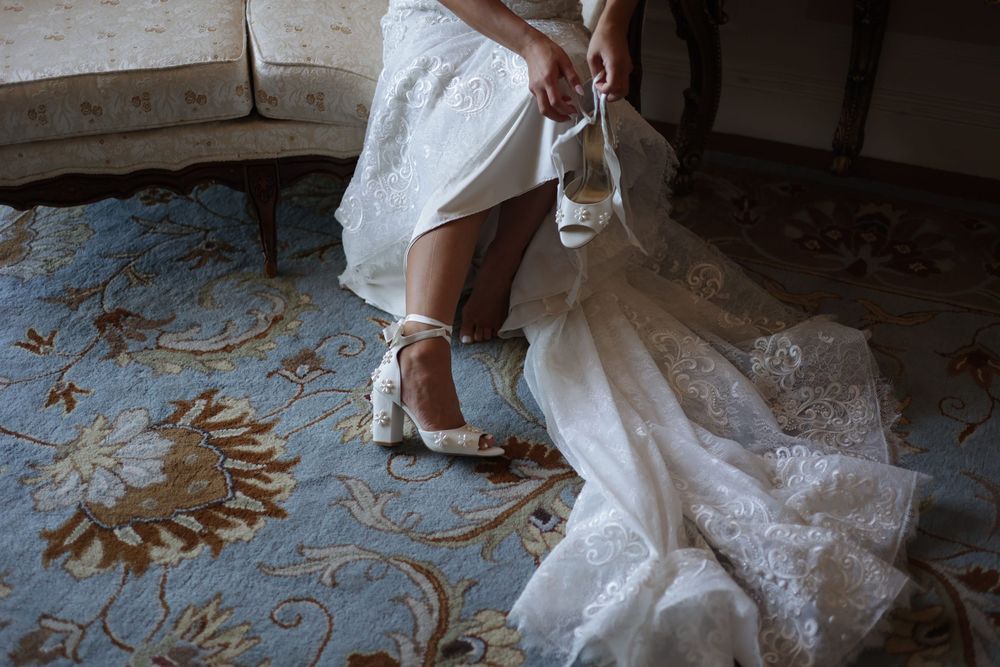 Bride in white lace wedding dress adjusting her elegant ankle-strap heels on a floral blue carpet.