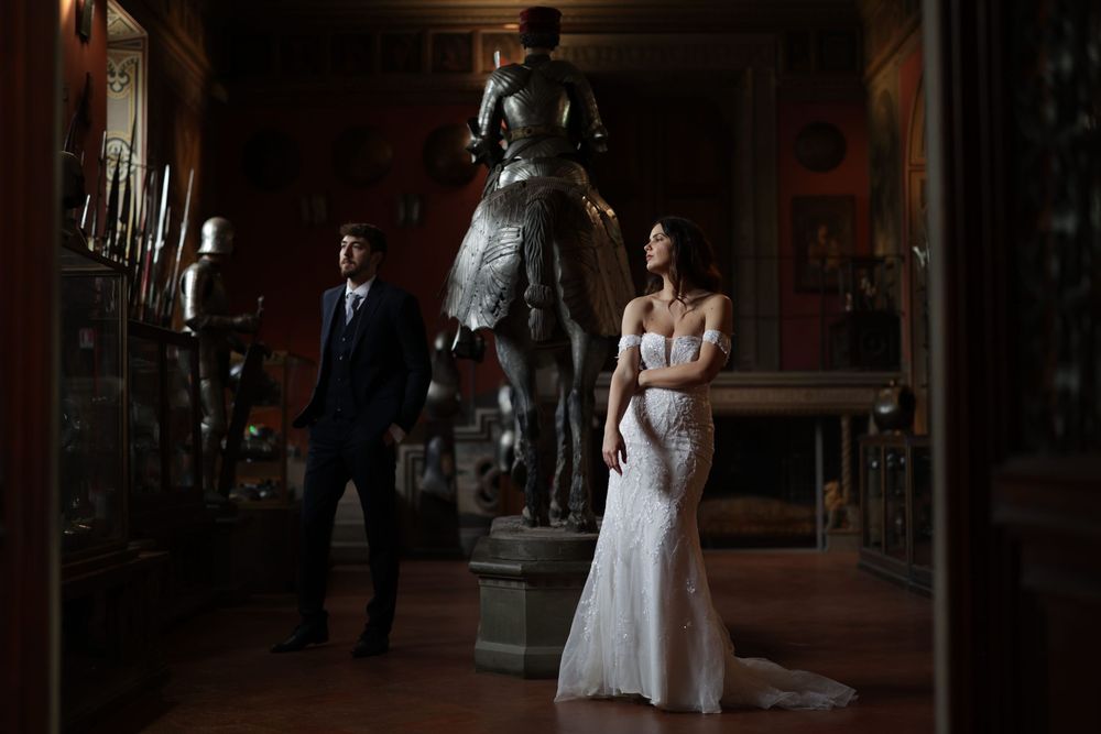 Bride in off-shoulder lace gown and groom stand beside medieval armor in a dimly lit castle hall.