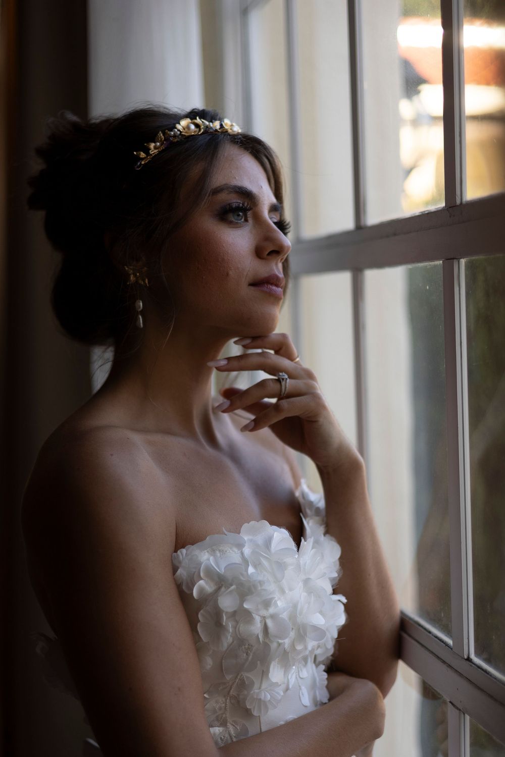 Bride in white floral strapless gown with gold tiara gazes thoughtfully out a window, hand delicately touching her chin.