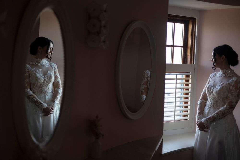 Bride in lace long-sleeve wedding gown admires her reflection in an oval mirror before her wedding ceremony.