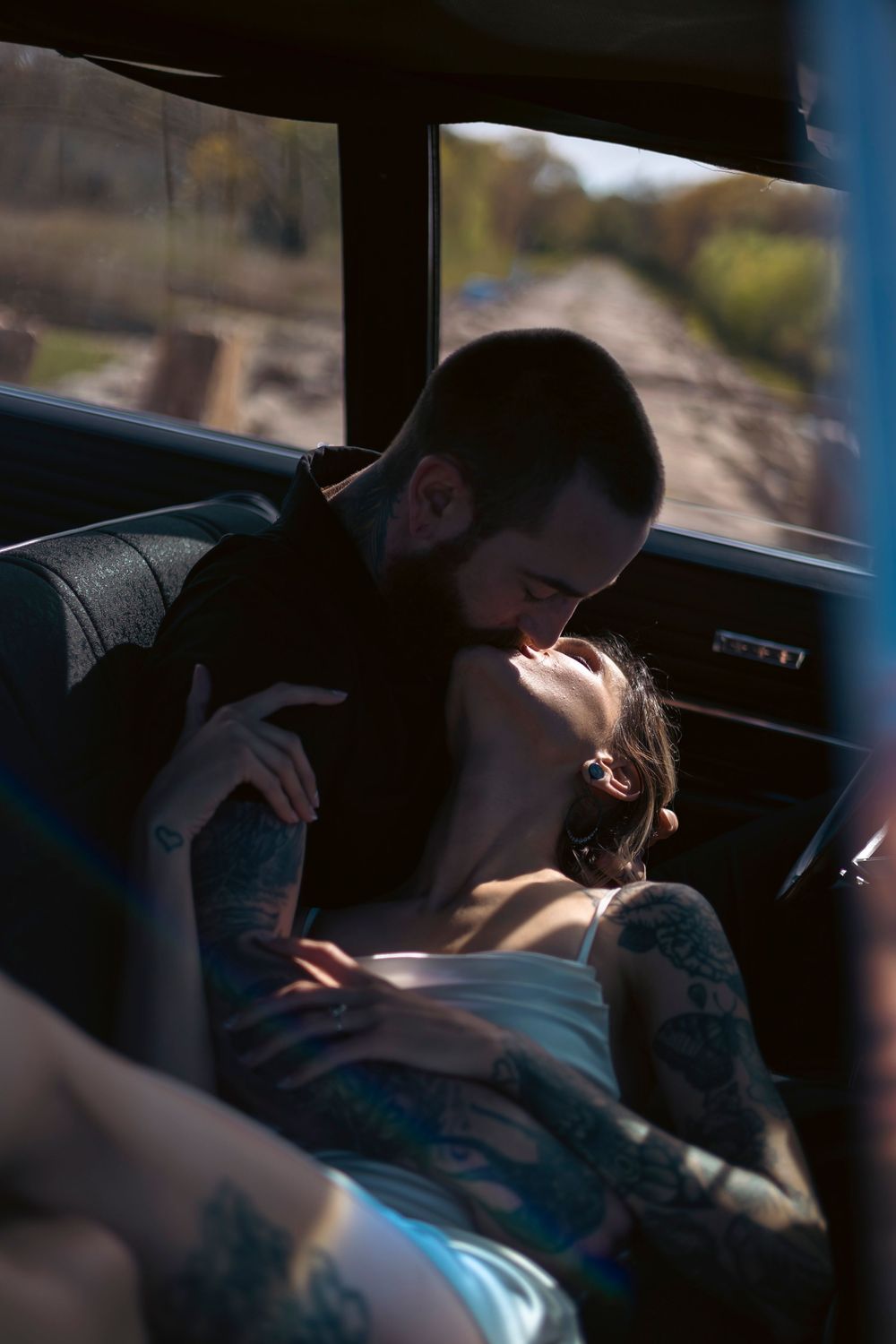 Tattooed couple sharing a romantic kiss inside a vintage car with sunlit countryside visible through the window.