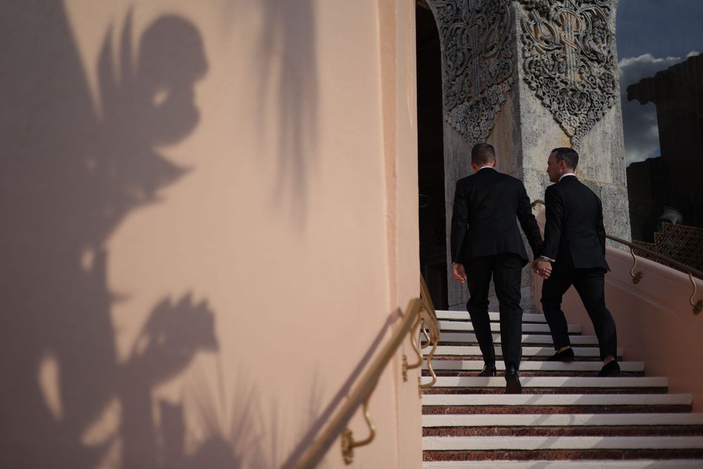 Two men in black suits walking up marble stairs into an elegant building with decorative stonework.