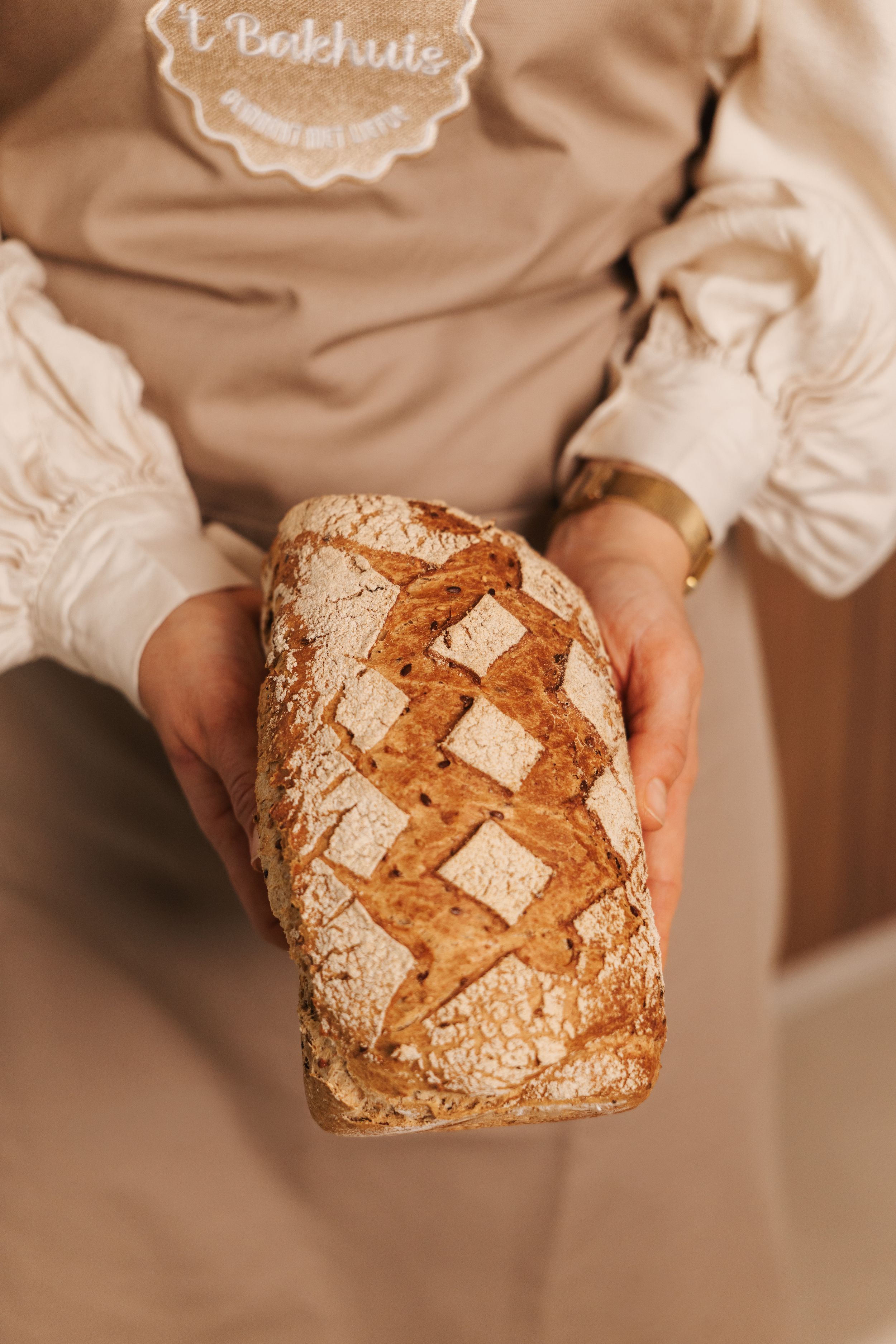 Baker holding a freshly baked artisan sourdough loaf with decorative scoring, wearing a 't Bakhuis apron.