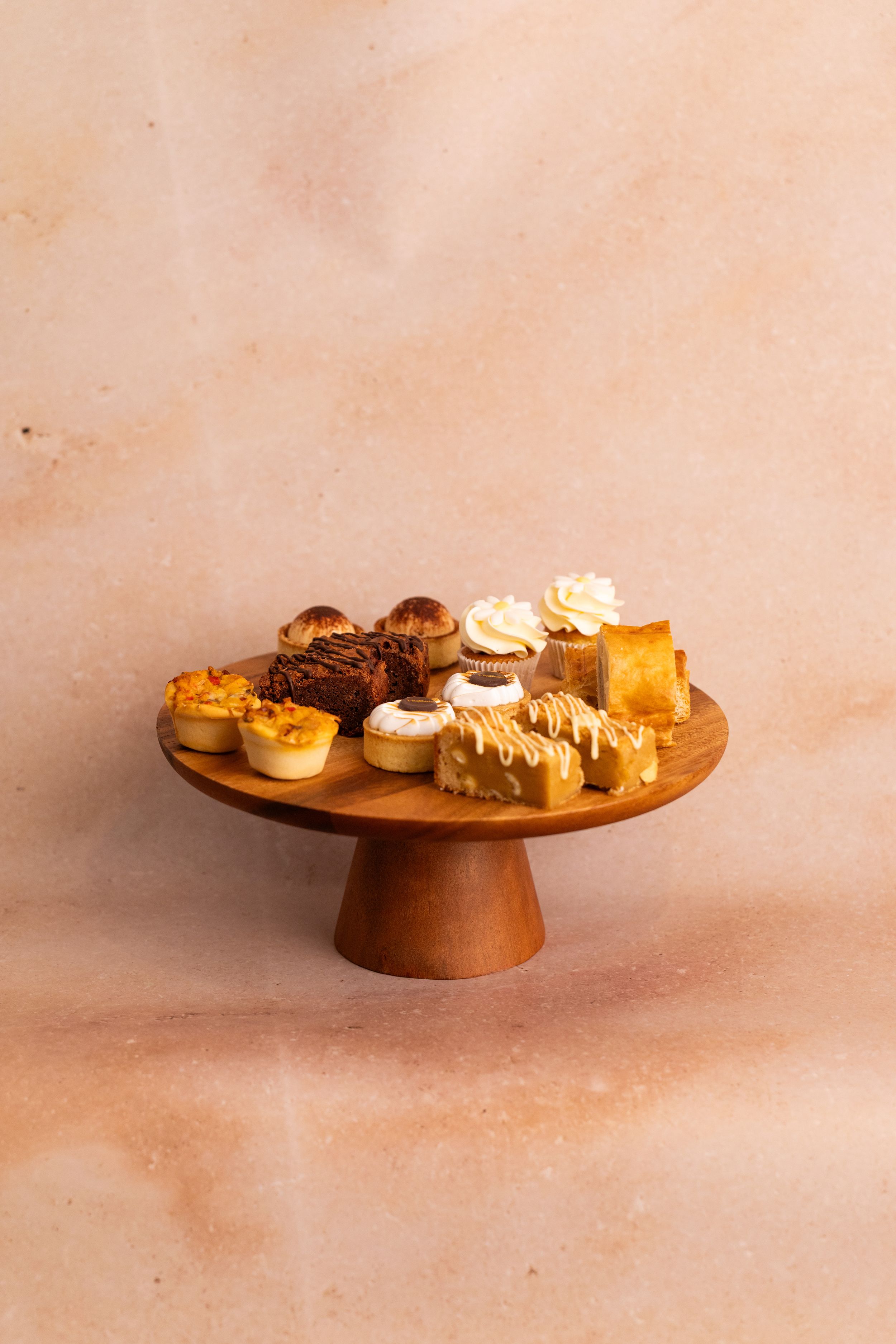 Assorted pastries and cupcakes displayed on a wooden cake stand against a warm beige background.