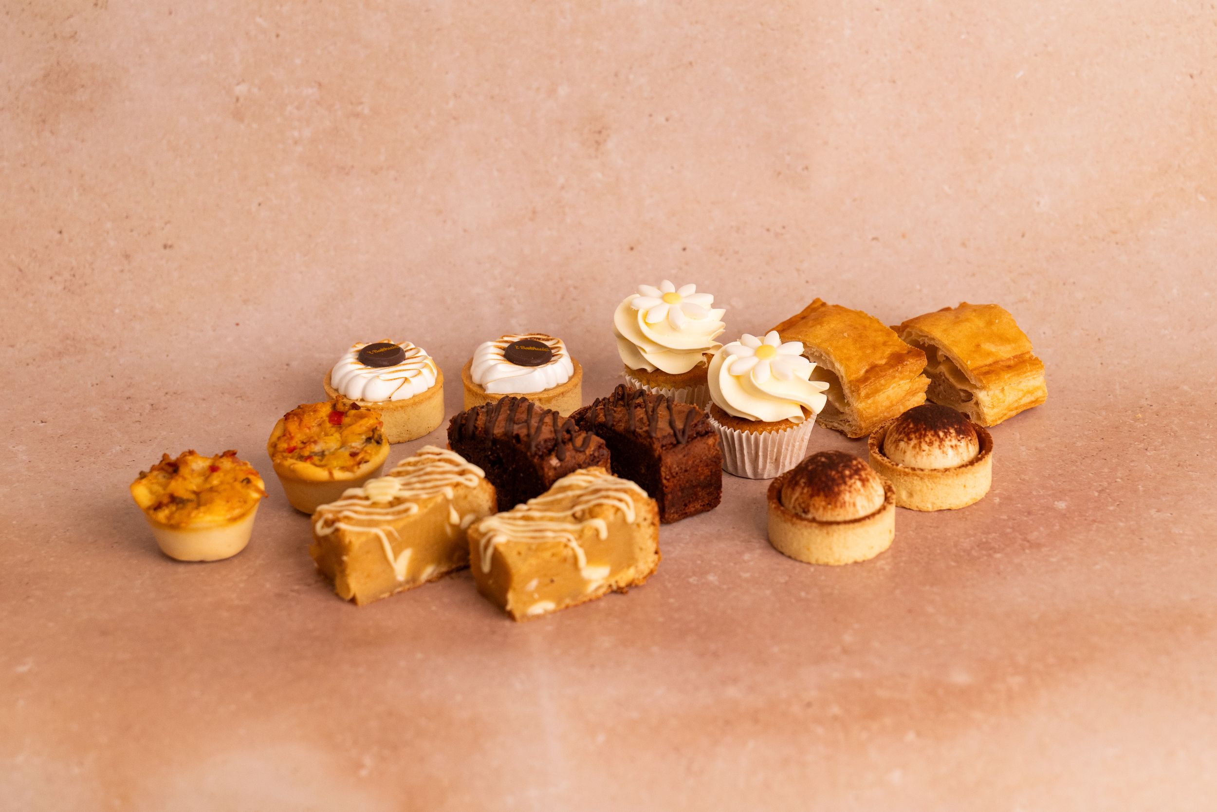 Assorted mini pastries and cupcakes including tarts, fudge, and frosted cupcakes displayed on a pink marble surface.