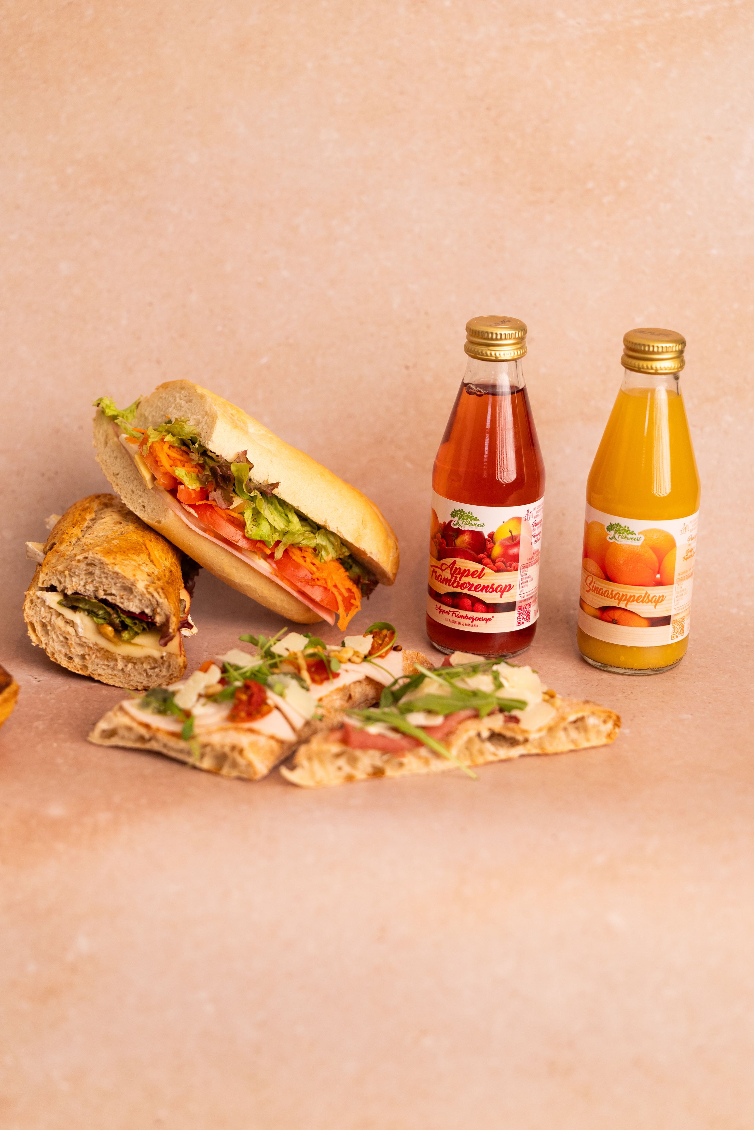 A sandwich, flatbread with toppings, and two juice bottles arranged on a beige surface.