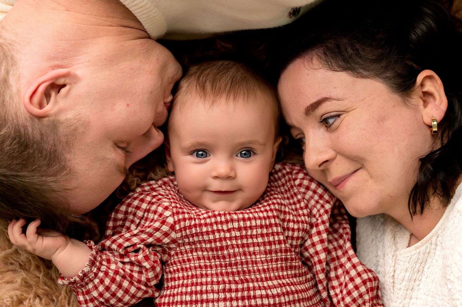 Two parents lovingly kiss their blue-eyed baby girl dressed in red gingham, captured in a tender family portrait.