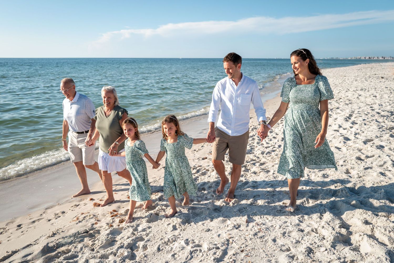 Multi-generational family walking on a sunny white sand beach, wearing matching green floral outfits, holding hands.